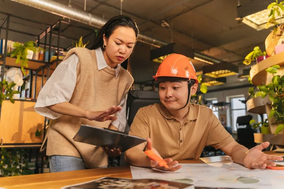 Two engineers discuss plans in an office, with one wearing a hard hat and the other holding a clipboard.
