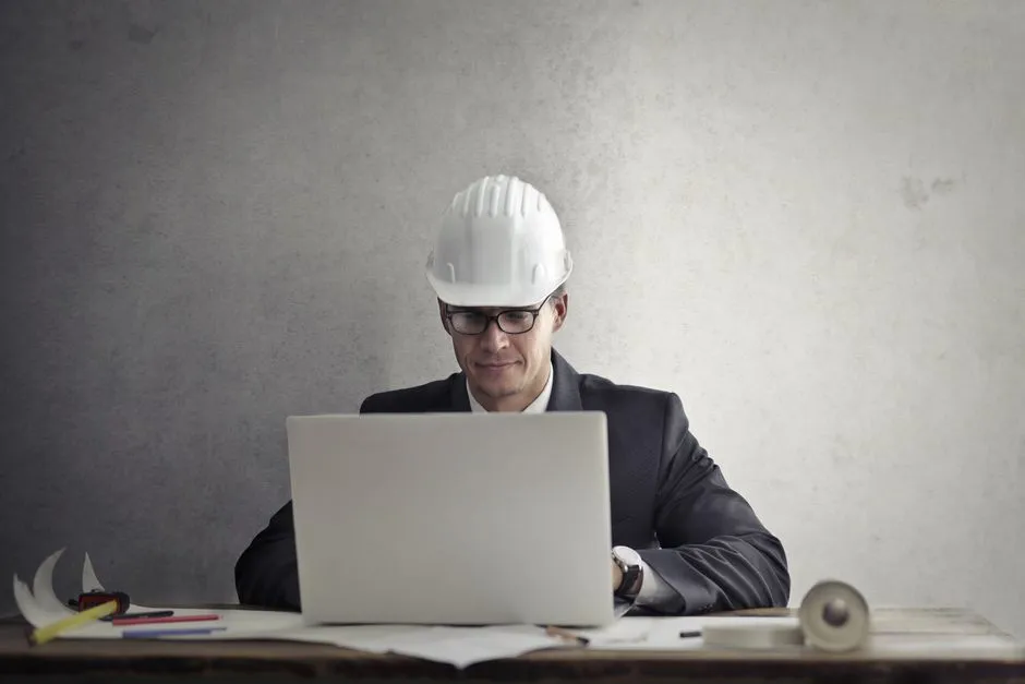 Serious professional male engineer in protective helmet and formal suit sitting at table and using laptop while working on project at workplace