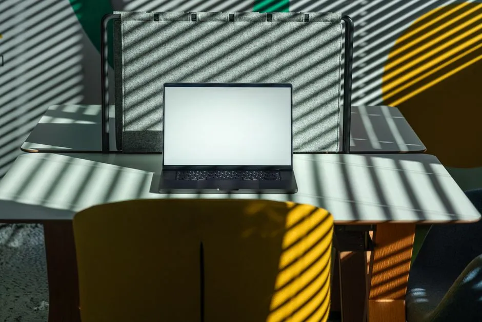 A modern office desk setup with a laptop, featuring geometric shadows from sunlight through blinds.