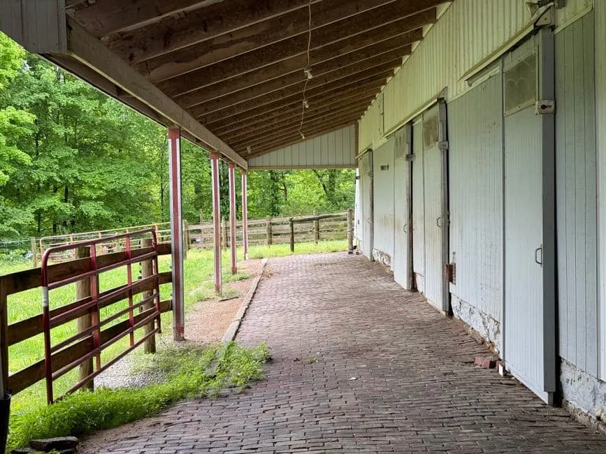 A serene barn aisle featuring brick flooring and exposed wooden beams, adjacent to lush greenery.
