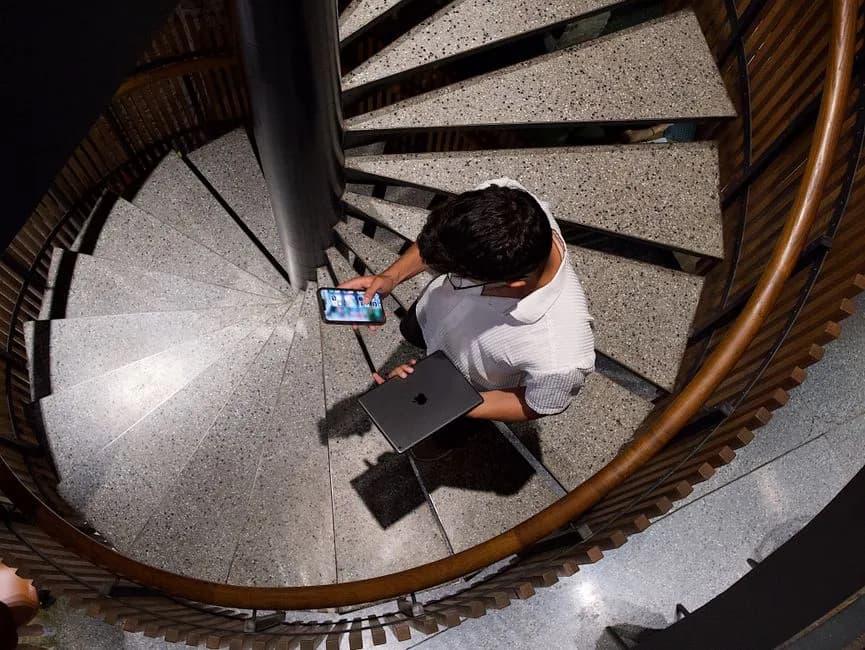 Top view of a man holding a smartphone and laptop on a spiral staircase indoors.