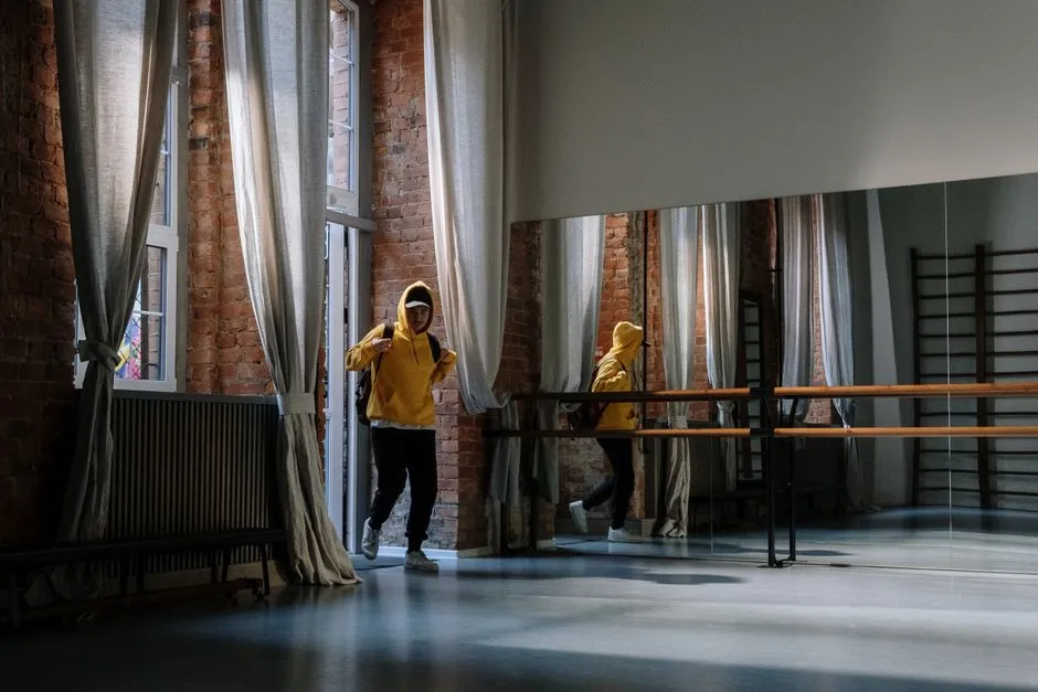 A young man in a yellow hoodie stands in a dance studio with a mirrored wall and brick interior.