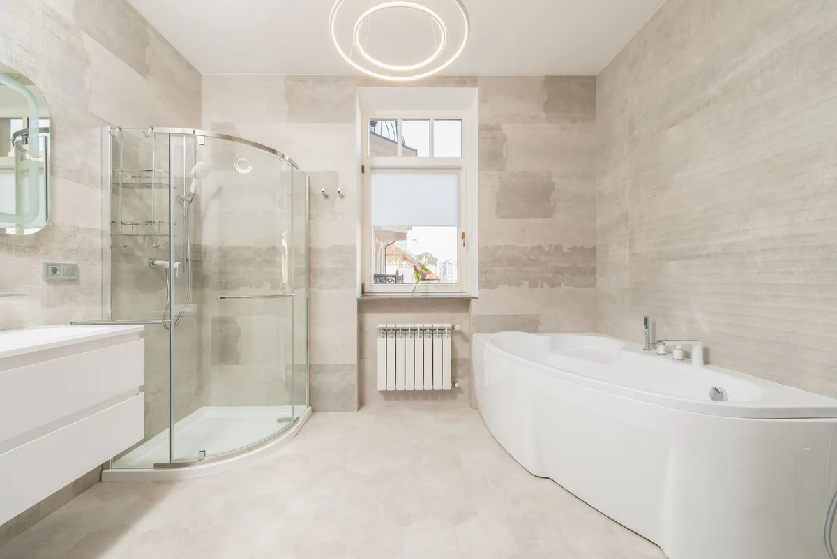 Finished light bathroom with a white bathtub and a glass shower enclosure near a window