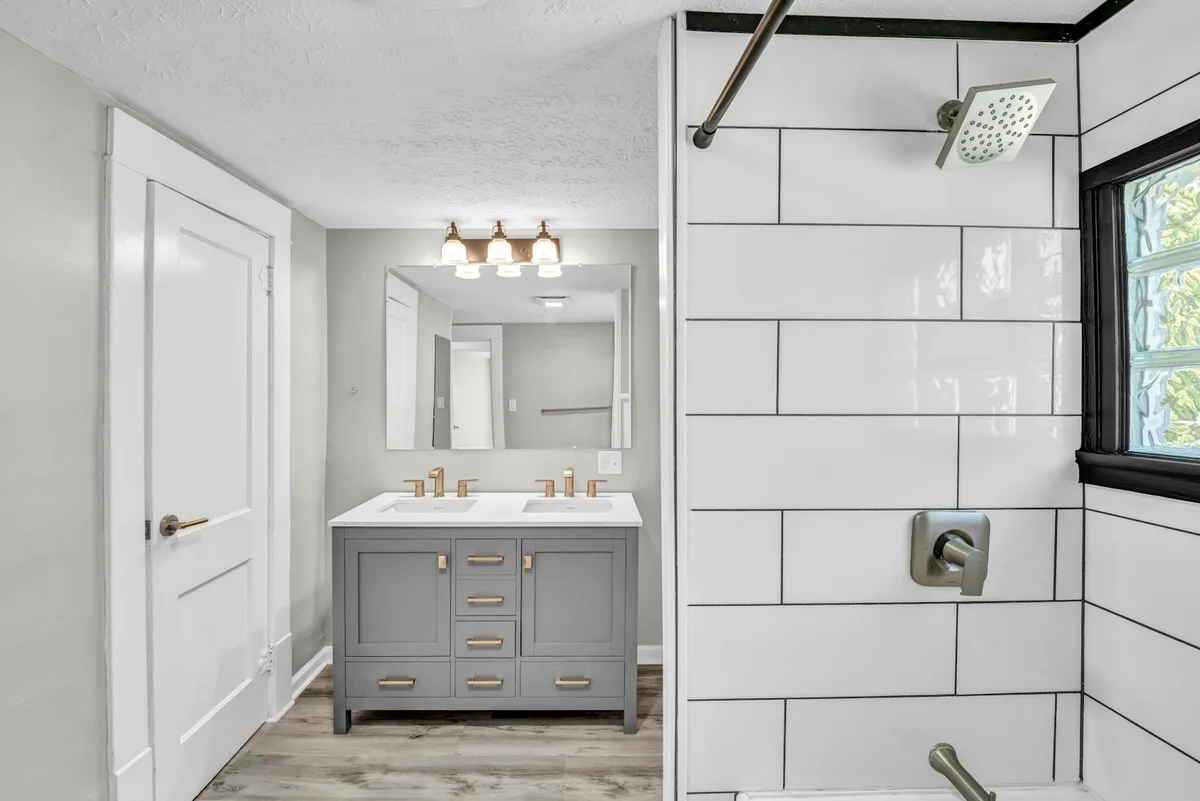 Bathroom with a grey double vanity, a shower lined with white subway tile and light wood flooring