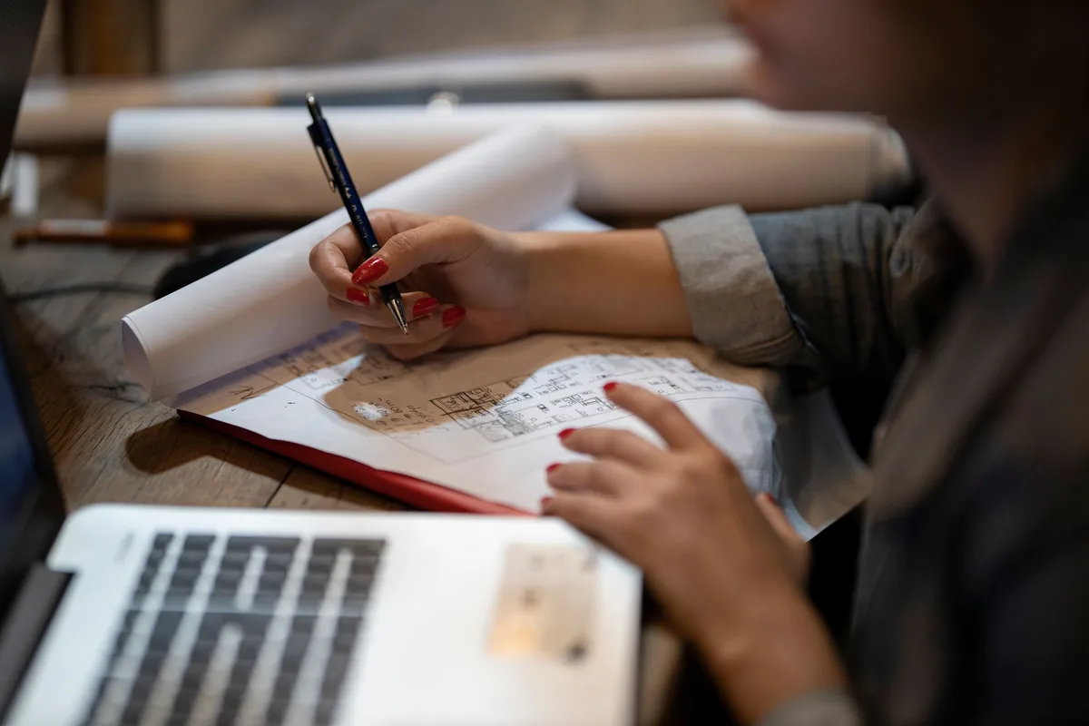 Person drawing architectural plans by hand next to a laptop at a desk